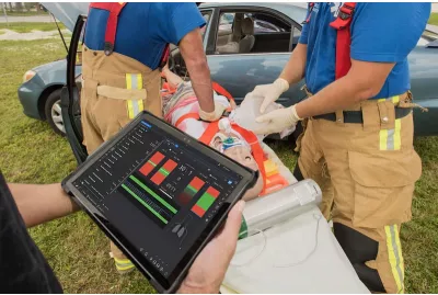 Trauma HAL patient simulator strapped to stretcher next to vehicle collision while two paramedics perform an assessment